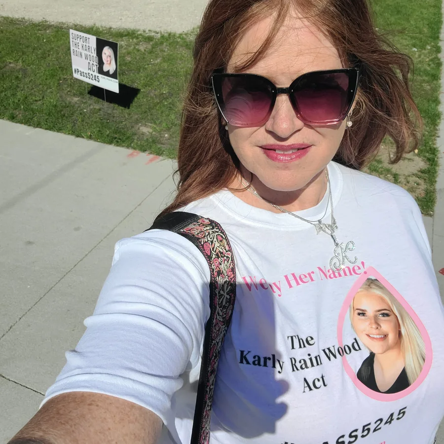 Amber Wood wearing a We Say Her Name shirt for the Karly Rain Wood Act, with a Support the Karly Rain Wood Act yard sign behind her.
