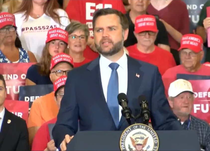Vice President JD Vance speaking at a public event. Behind him in the crowd, a supporter wears a white #PASS5245 Karly Rain Wood Act shirt, circled with a pink heart.