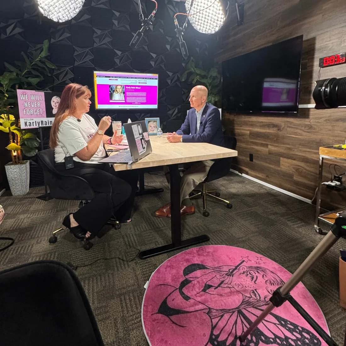 A podcast interview in progress with Senator Pete Ricketts, with a Karly Rain Wood page on the studio screen and a We Will Never Forget sign.